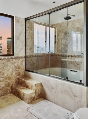A bathroom with a glass-enclosed shower, marble walls, and a view of the cityscape.
