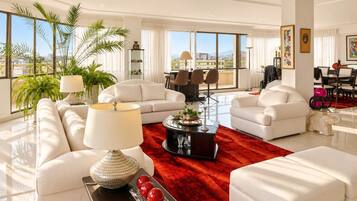 A spacious living room with white sofas, a red rug, and a large window offering a city view.