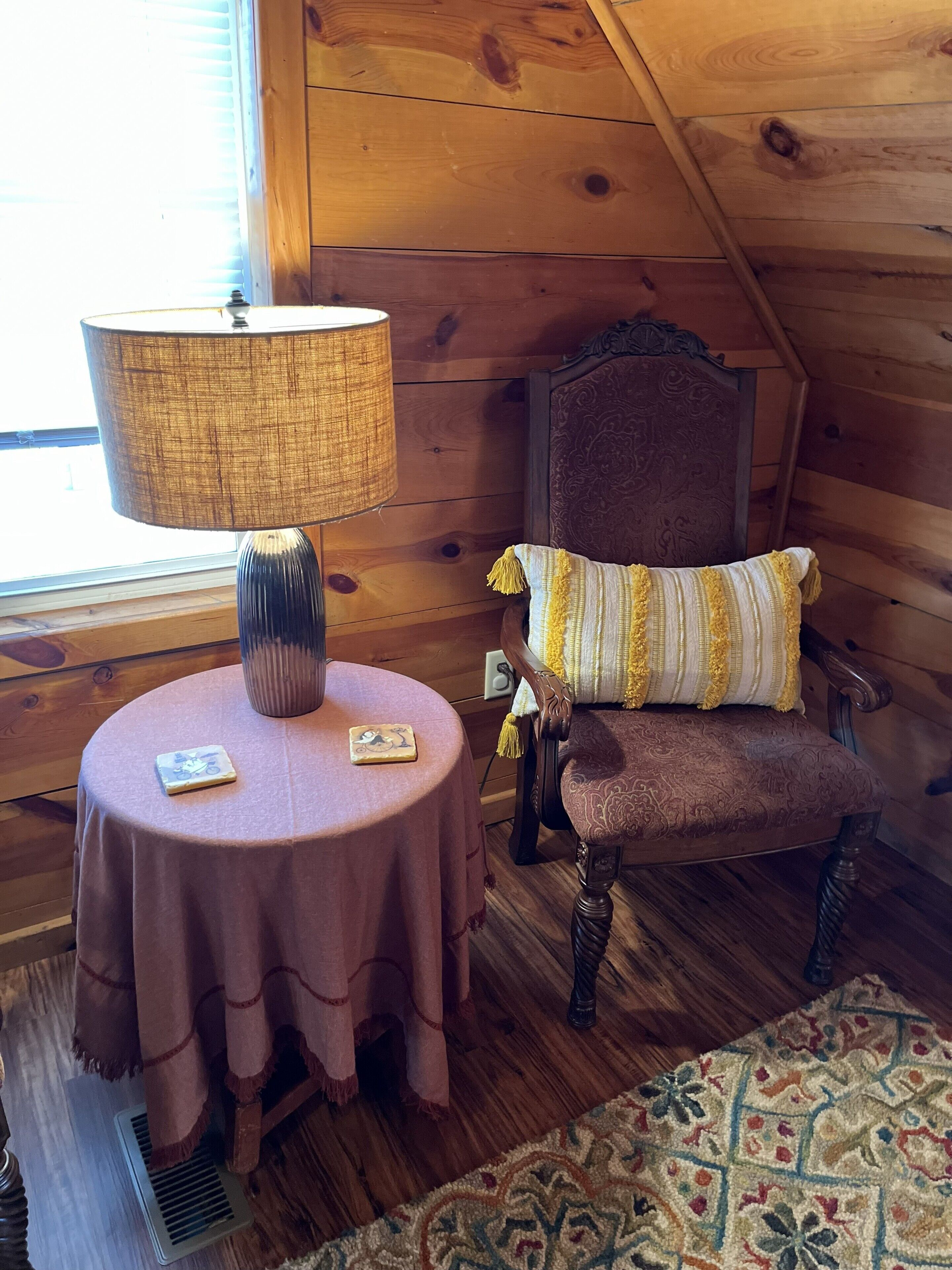 A cozy room with a round table, a lamp, a chair with a patterned cushion, and a patterned rug.