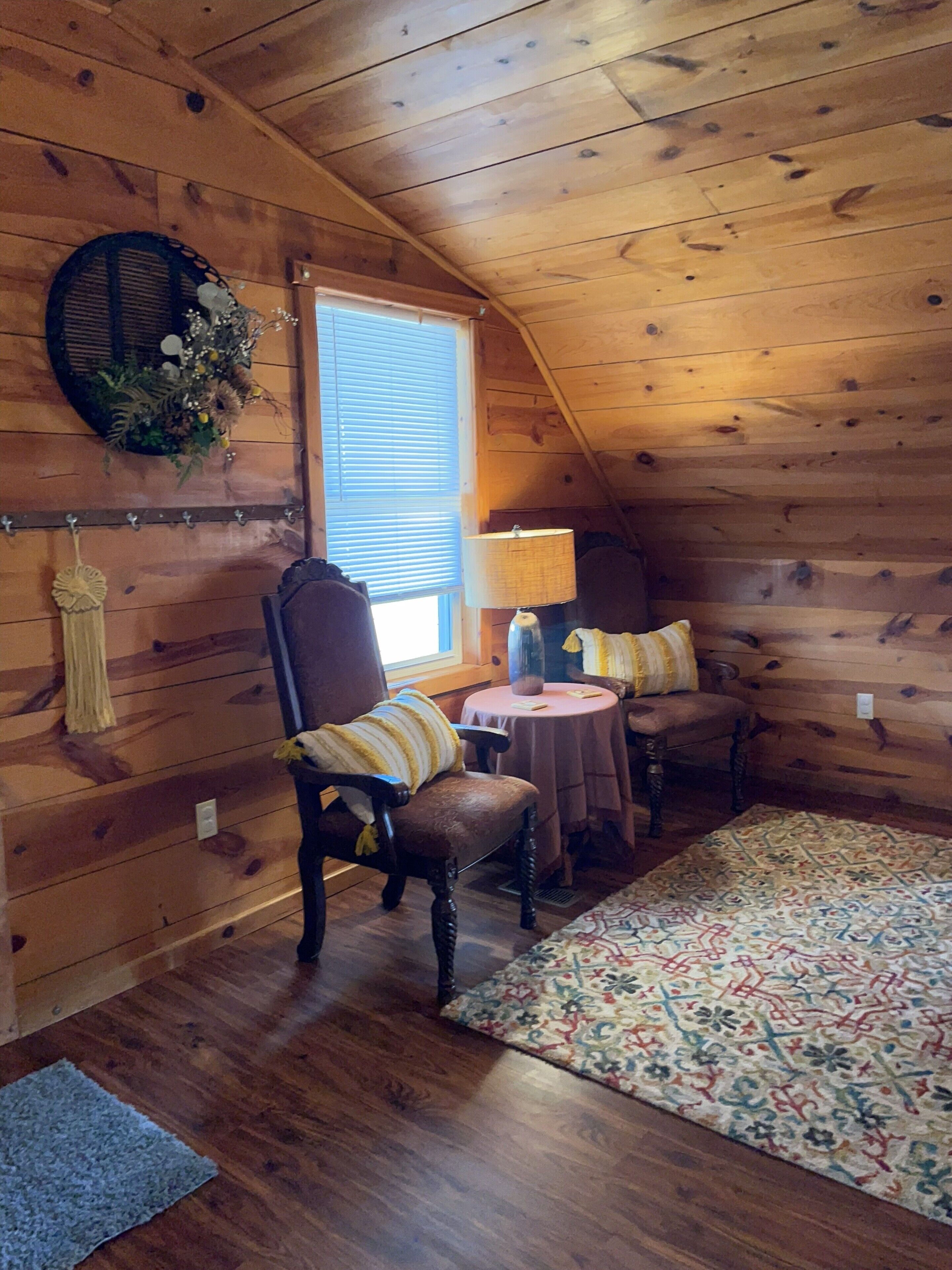 Acogedor dormitorio en ático con paredes de madera, una ventana, una lámpara, una silla, una mesa y una alfombra estampada.