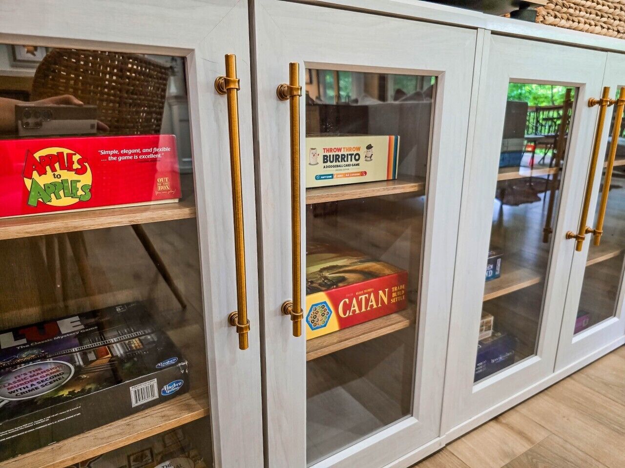 Ein Schrank mit Glastüren und Messinggriffen, der Gesellschaftsspiele wie Apples to Apes, Throw Throw Burrito und Catan beherbergt.