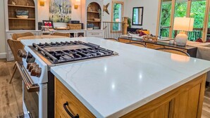 A modern kitchen with a large island, built-in oven, and gas stove. There are wicker chairs and a dining table in the background.