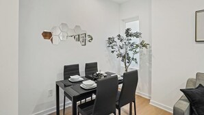 Un coin repas avec une table dressée pour quatre personnes, des chaises noires, un miroir hexagonal et une plante en pot.