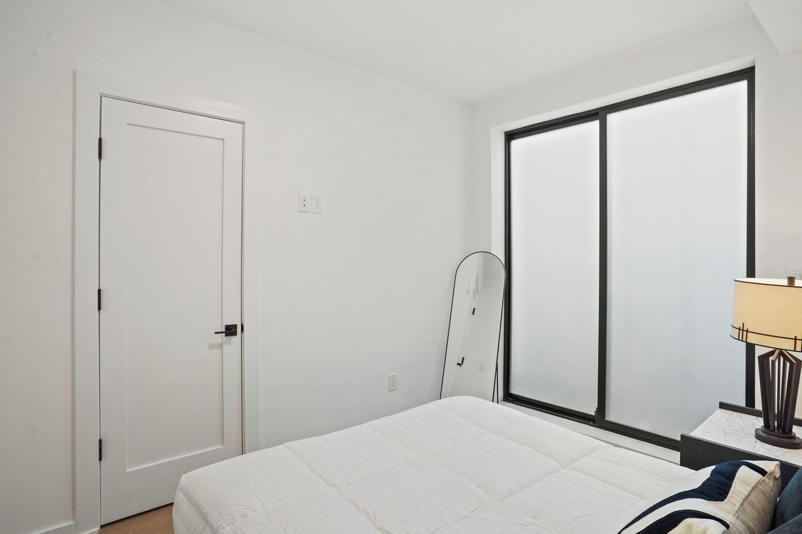 Ein Schlafzimmer mit einem Bett, einem Fenster, einer Tür und einem Nachttisch mit einer Lampe.