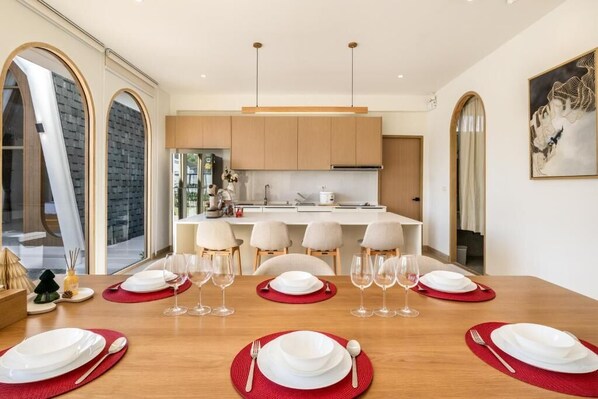 A modern kitchen with a central island, dining table set for six with red placemats, and large windows with arched frames. - Winvilla Pattaya (Bang Lamung)