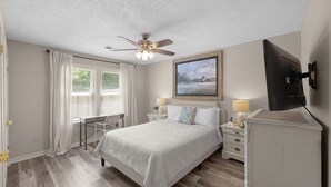 Quarto com cama, mesas de cabeceira, ventilador de teto, televisão e janela com cortinas.