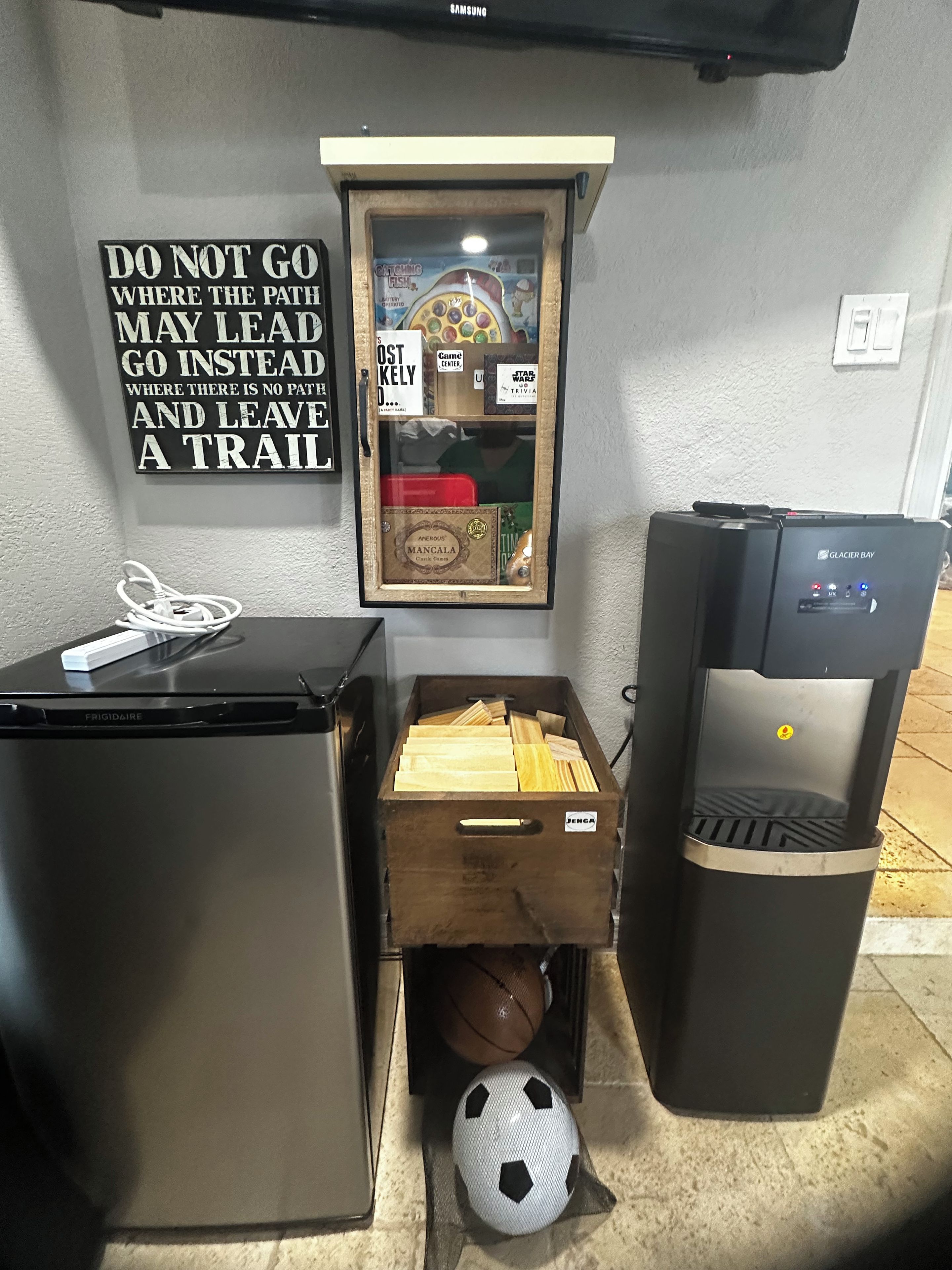 A kitchenette with a refrigerator, a drawer with wooden planks, a water dispenser, and a wall with a motivational quote.