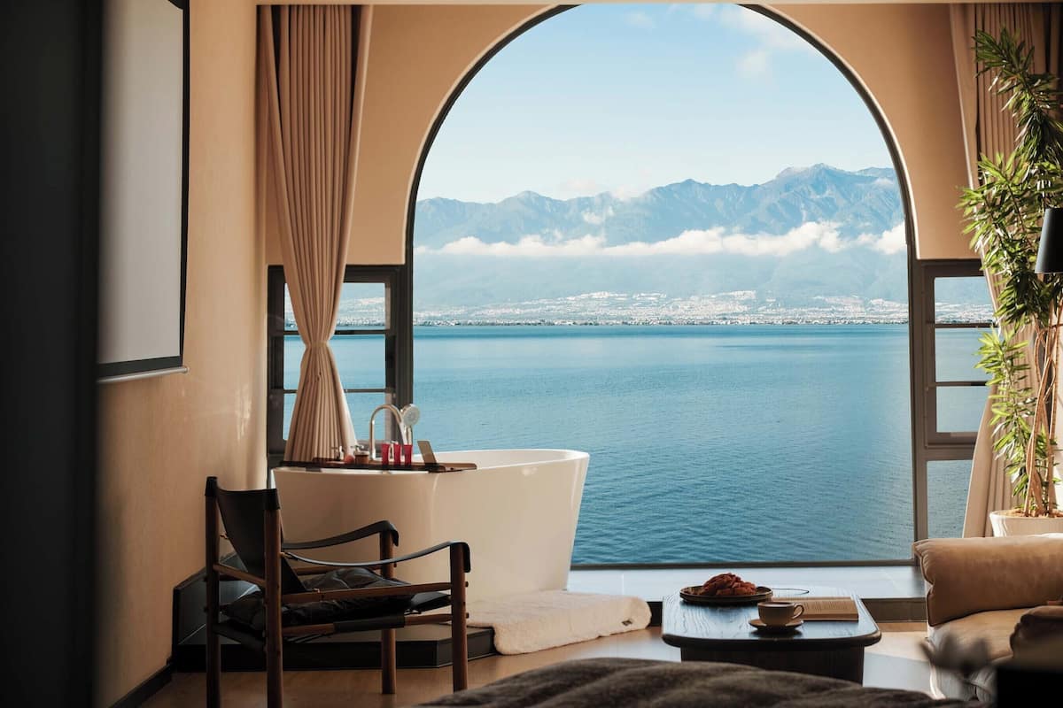 A room with a large arched window, a bathtub, a chair, a small table with a bowl, and a potted plant.