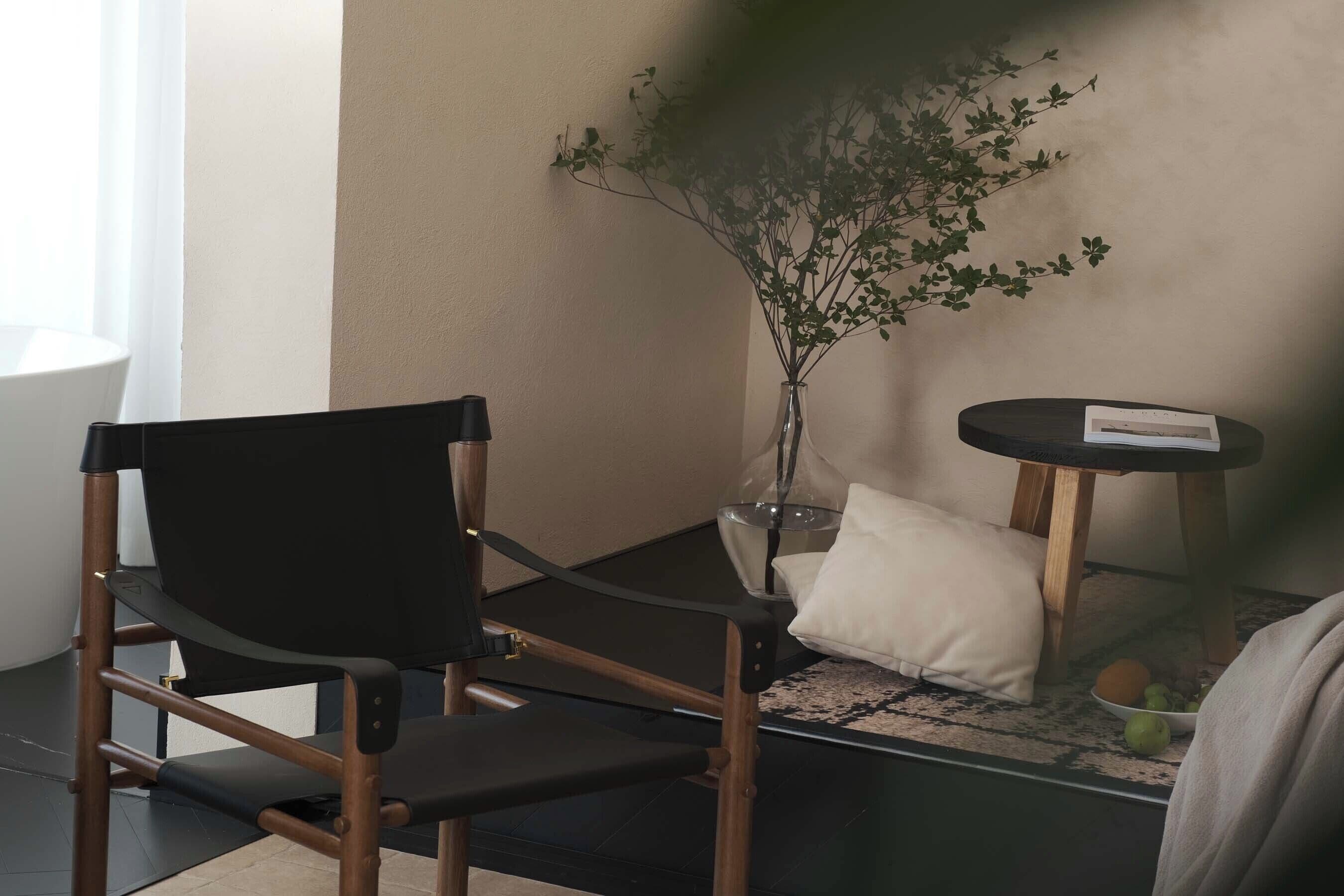 A room with a black chair, a small table with a book, a vase with a plant, and a cushion.
