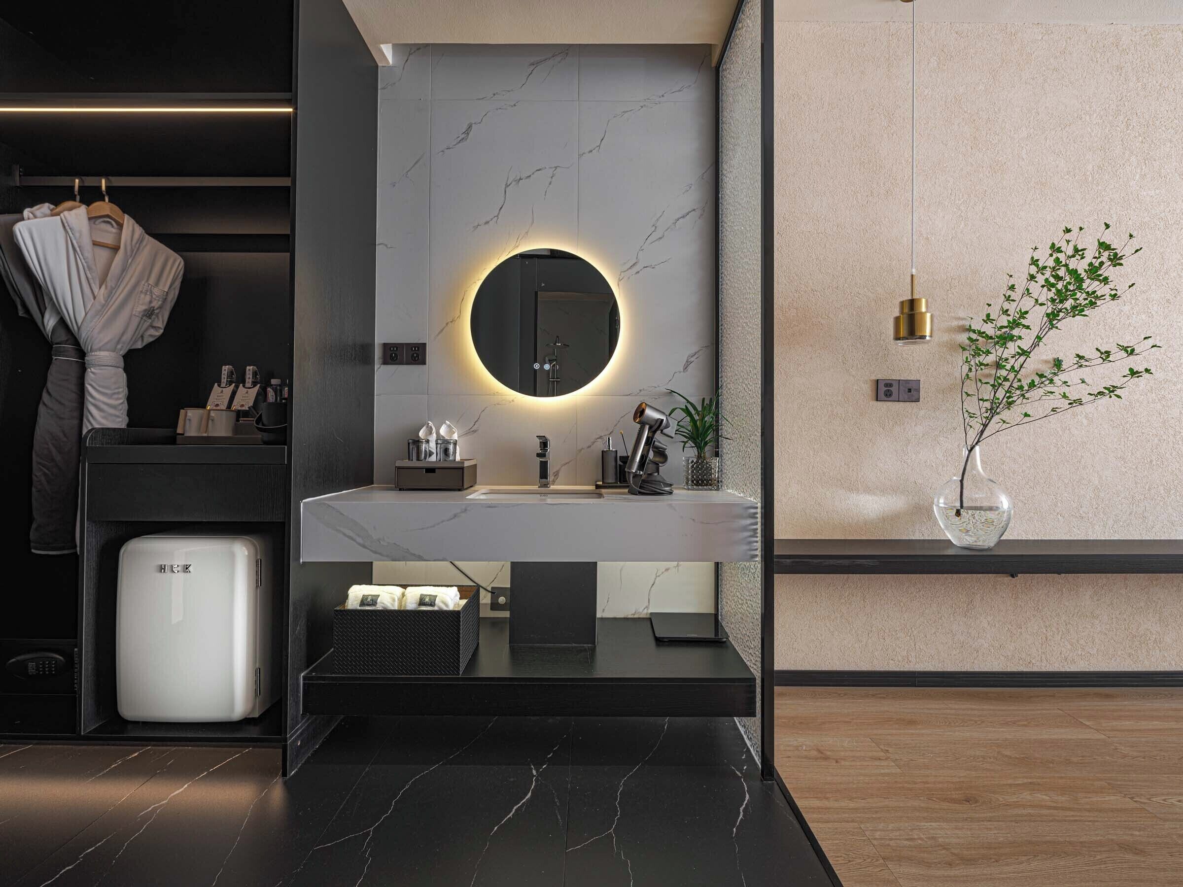 A modern bathroom with a marble vanity, a round mirror with a light, a sink, a towel rack, and a small trash bin.