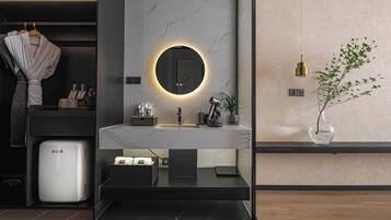 A modern bathroom with a marble vanity, a round mirror with a light, a sink, a towel rack, and a small trash bin.