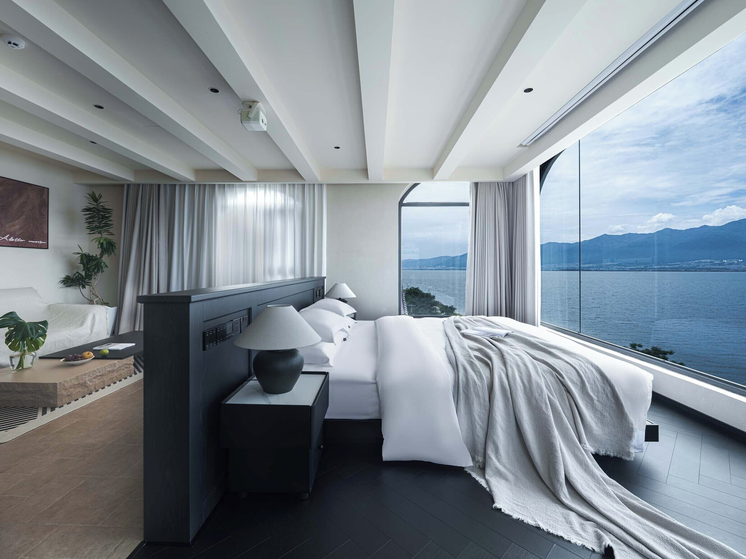 A modern hotel room with a large window overlooking a body of water, a bed with white linens, a bedside table with a lamp, and a small plant on the side table.