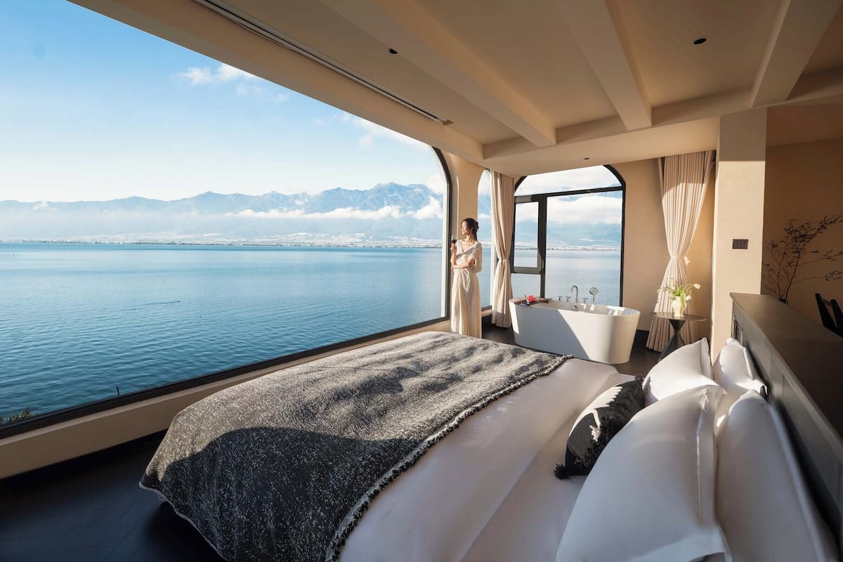 A bedroom with a large bed, a bathtub, and a view of a body of water and mountains.