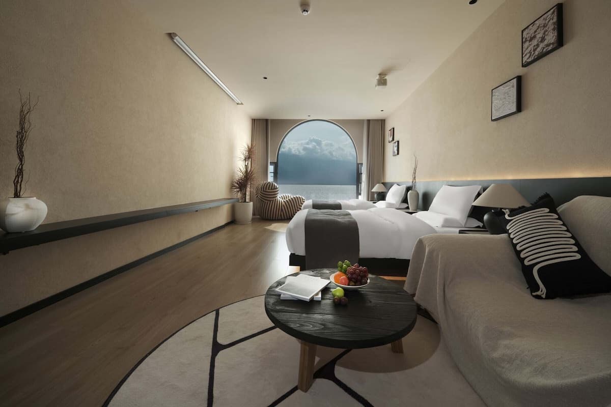 A hotel room with a bed, a sofa, a small table with fruit, a potted plant, and a view of the sea.