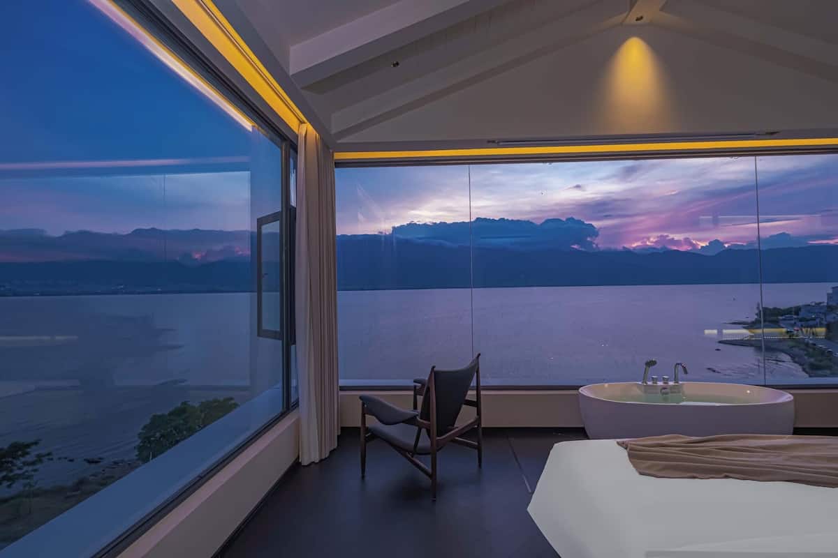A modern hotel room with a large window, a bed, a chair, and a round table with a sink.