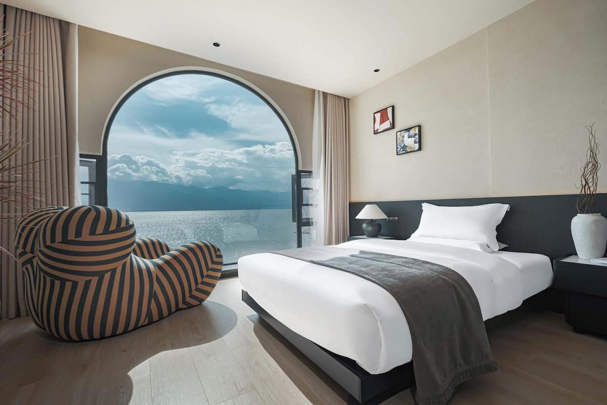 A modern hotel room with a large bed, a curved armchair, a nightstand, a lamp, and a view of the sea through a large window.