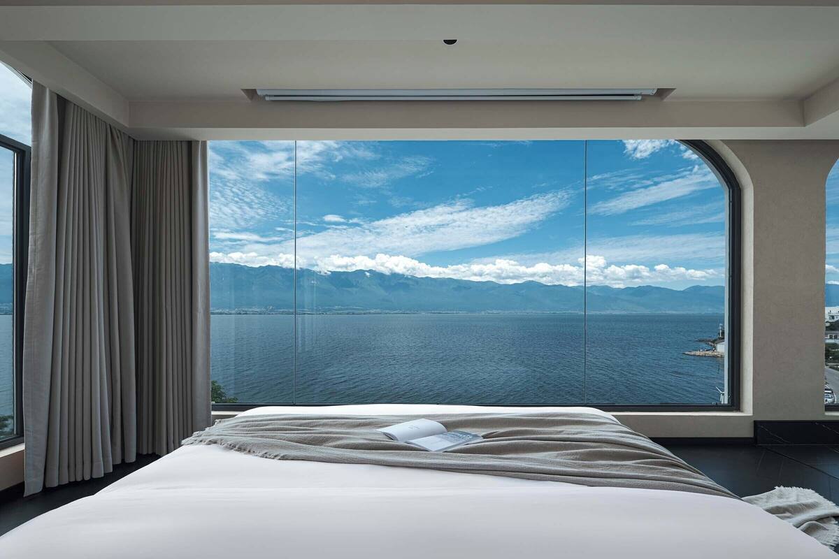 A bedroom with a large window, a bed, and a view of the sea and mountains.