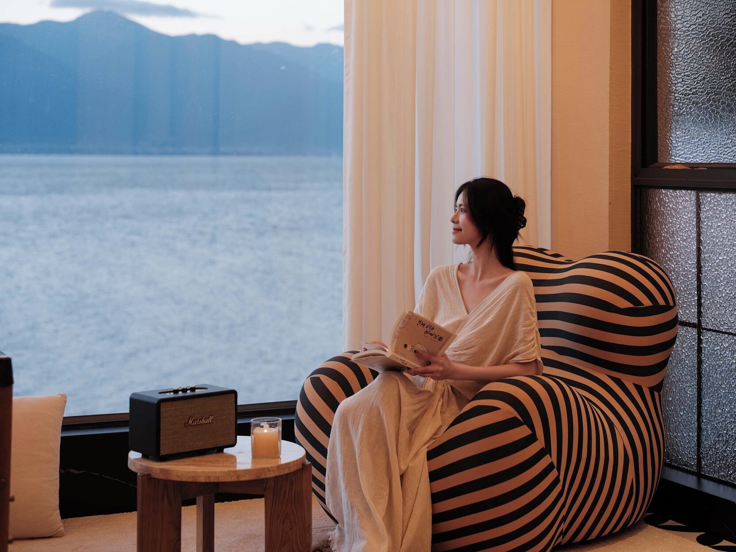 A person sitting on a striped bean bag chair by a window with a view of a body of water and mountains.