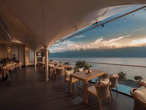 An outdoor dining area with tables and chairs, a view of the sea, and a sunset.