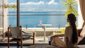 A room with a view of a body of water and mountains, a chair, a table with drinks, and a person sitting by the window. - Dali Azure Hillside Boutique Resort- Wenbi Village - Gourmet Dining & Cocktail Lounge & Complimentary Airport Pick-up for 2 Nights (Dali)