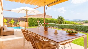 Eine Terrasse im Freien mit einem Holztisch, Korbstühlen, einem weißen Sonnenschirm und einem Pool.
