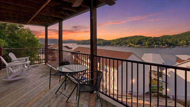 Una terraza de madera con una mesa y sillas, con vista a un lago y colinas.