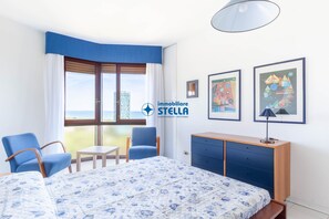 Ein Hotelzimmer mit einem Bett, zwei Stühlen, einem kleinen Tisch, einem Schrank und Blick aufs Meer.