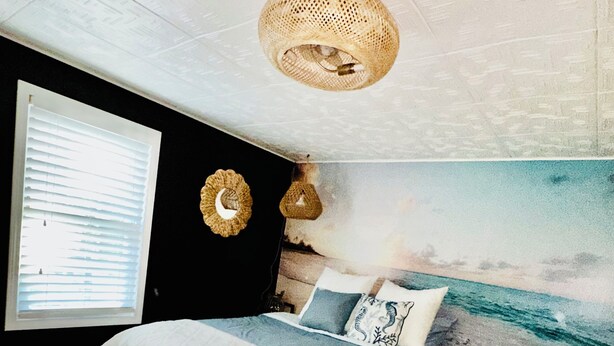 A bedroom with a bed, a wall clock, a window with blinds, a beach mural, and a ceiling light fixture.