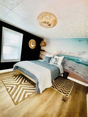 A bedroom with a bed, a wall clock, a window with blinds, a beach mural, and a ceiling light fixture.