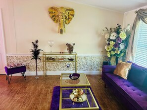 A living room with a purple sofa, a glass coffee table, a decorative elephant wall piece, a floral arrangement, and a marble-patterned wall.