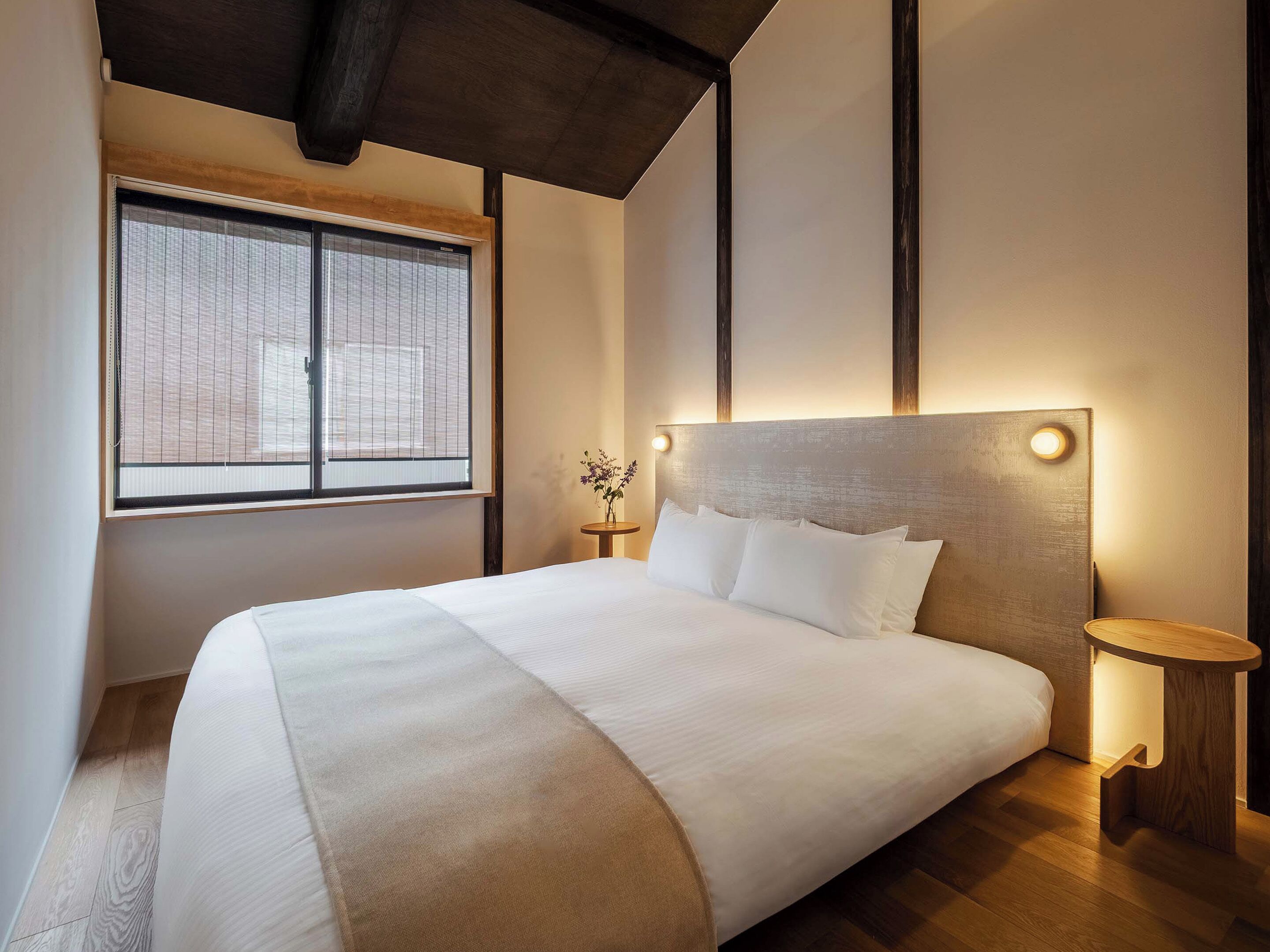 A modern hotel room with a large bed, a small bedside table, a nightstand, a window with blinds, and a wooden ceiling.