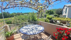 Ein Balkon mit rundem Tisch und Stühlen, umgeben von Kübelpflanzen und Blick auf eine üppige Landschaft.