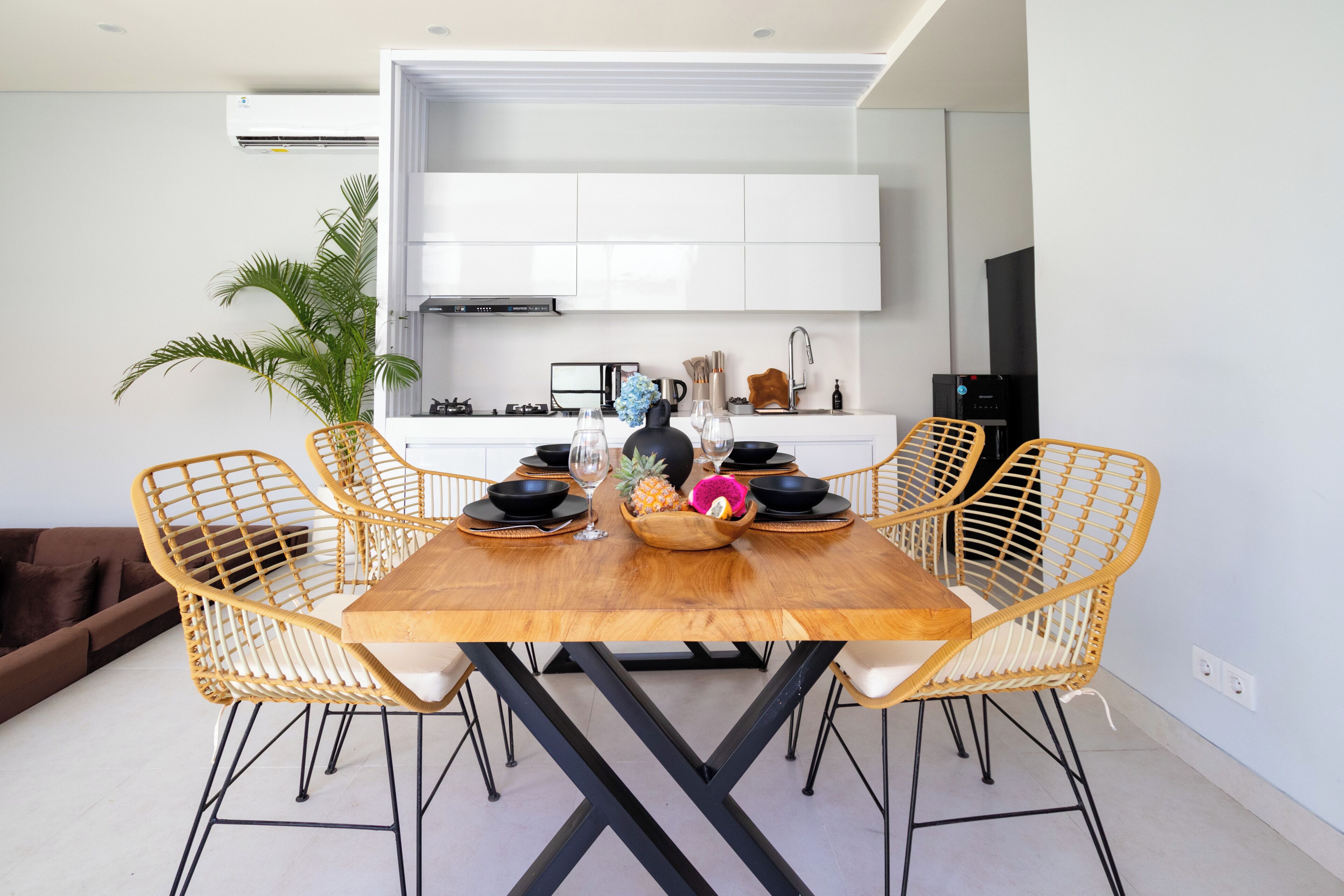 Un coin repas avec une table en bois, quatre chaises en osier, un grand saladier rempli de fruits au centre, et une cuisine en arrière-plan.