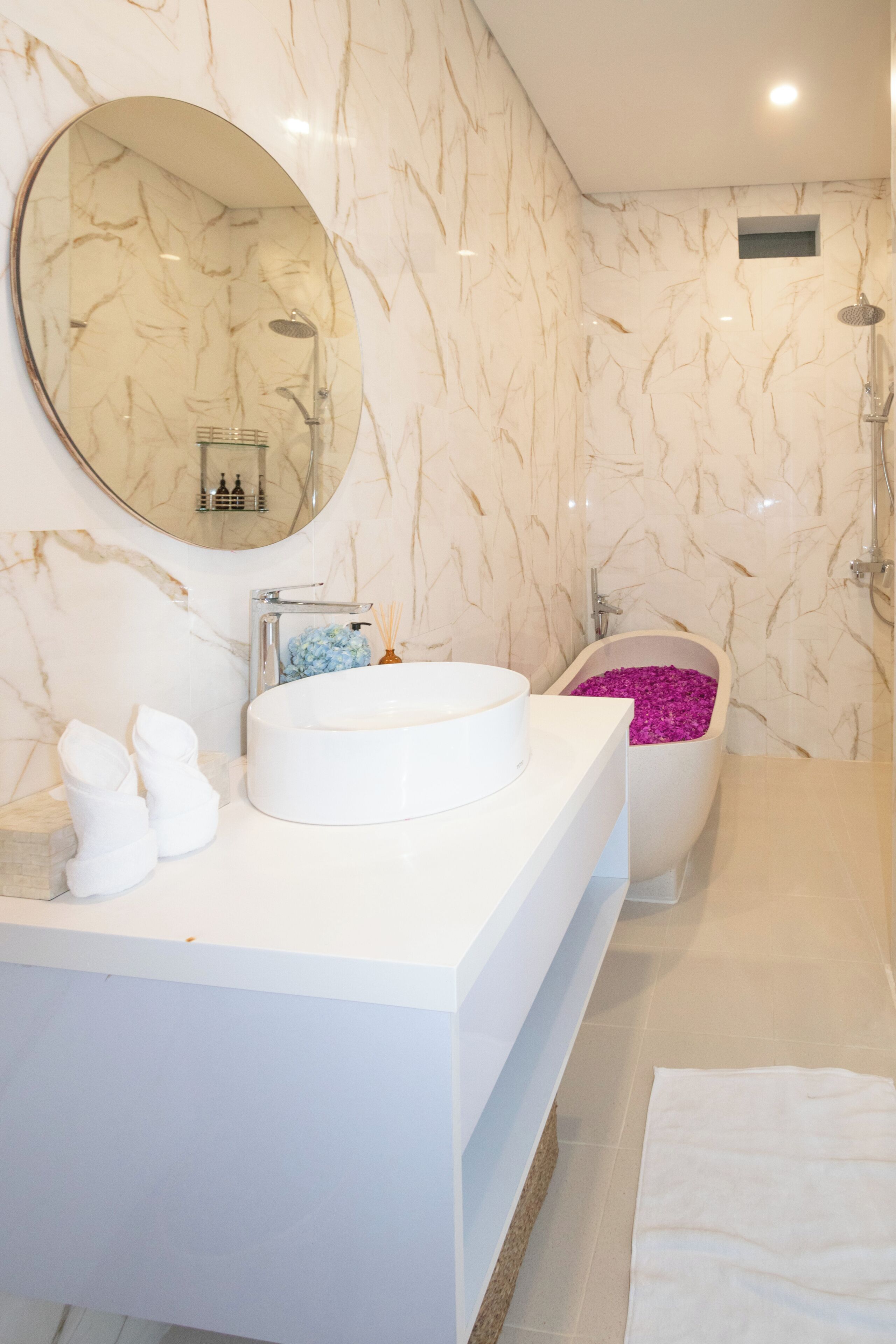 A bathroom with a marble wall, a round mirror, a white sink, a bathtub with pink flowers, and a shower area.