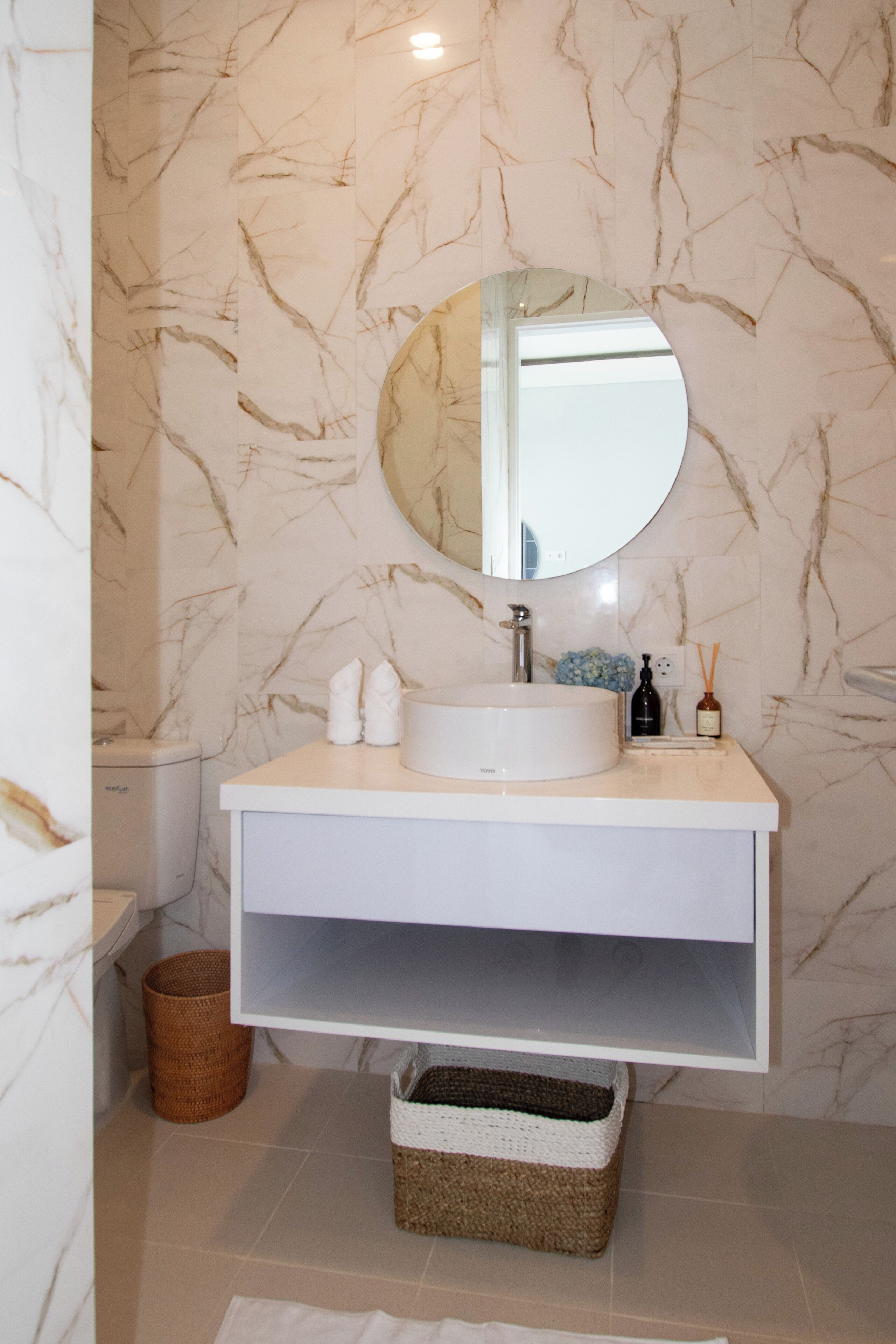A bathroom with a marble wall, a round mirror, a white sink, and a storage basket.