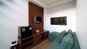 Een knus woongedeelte met een flatscreen televisie aan een houten wandpaneel, een kleine zwarte koelkast, een groene bank en een afvalbak.