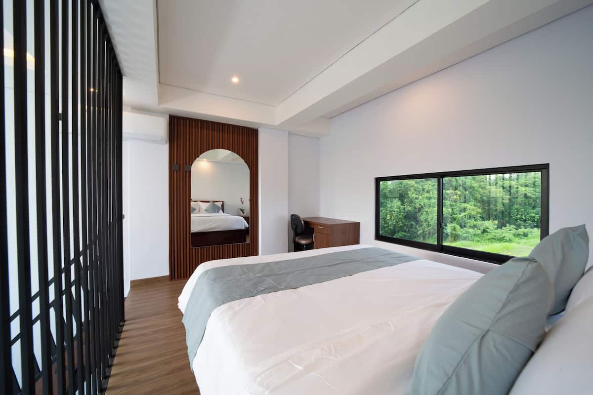A modern bedroom with a large bed, a desk, and a window with a forest view.