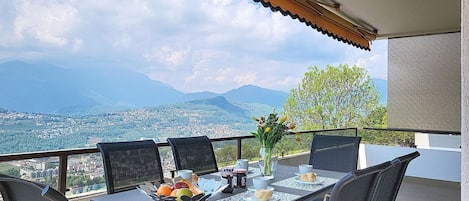 A balcony with a table set for two, a vase with flowers, and a scenic view of a city and mountains.