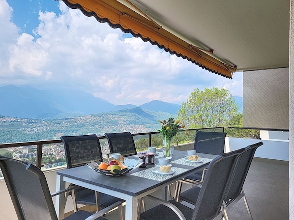 Ein Balkon mit Tisch für zwei Personen, einer Blumenvase und einem weiten Blick auf Stadt und Berge.
