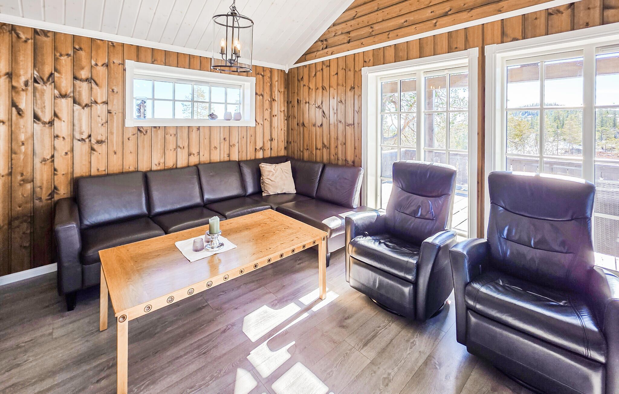 A room with a wooden paneled wall, a black leather sofa, two leather armchairs, a wooden coffee table, and a chandelier.