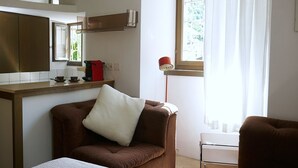 Une pièce avec un fauteuil marron, une petite table sur laquelle se trouve une lampe rouge, une table de chevet blanche et une fenêtre avec des rideaux.