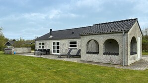 Un bâtiment en pierre avec un toit en tuiles, une terrasse avec des chaises et une table, et une pelouse verdoyante.