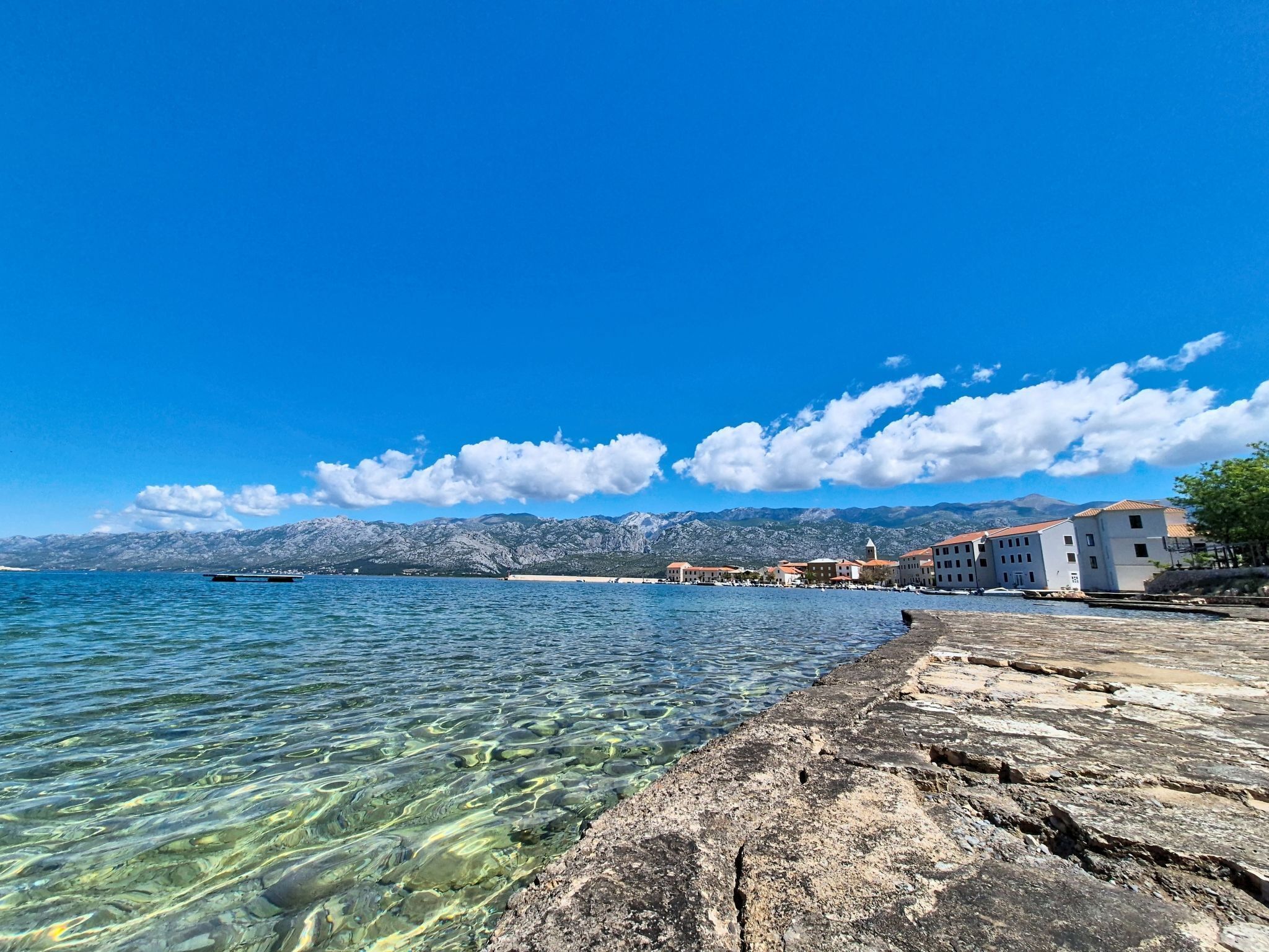 A coastal town with clear waters, rocky shoreline, and mountains in the background.