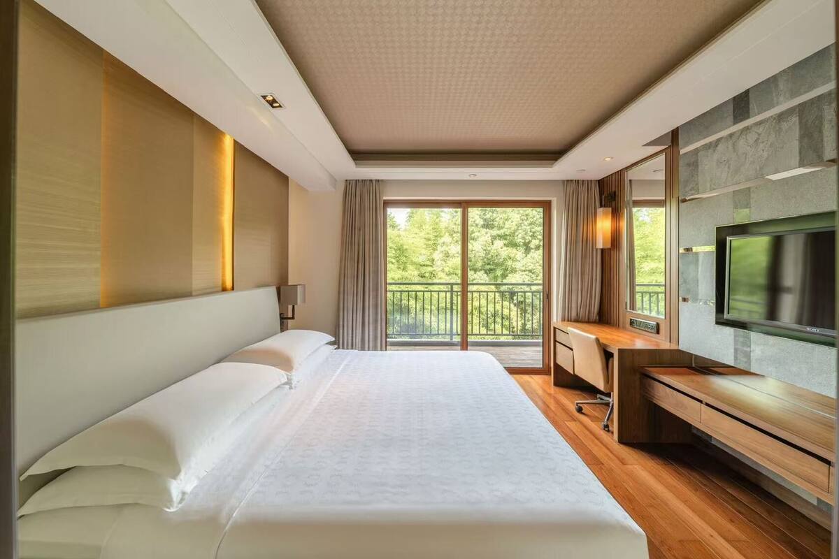 Una habitación de hotel moderna con una cama grande, un escritorio, un televisor de pantalla plana y un balcón con vistas a la vegetación.