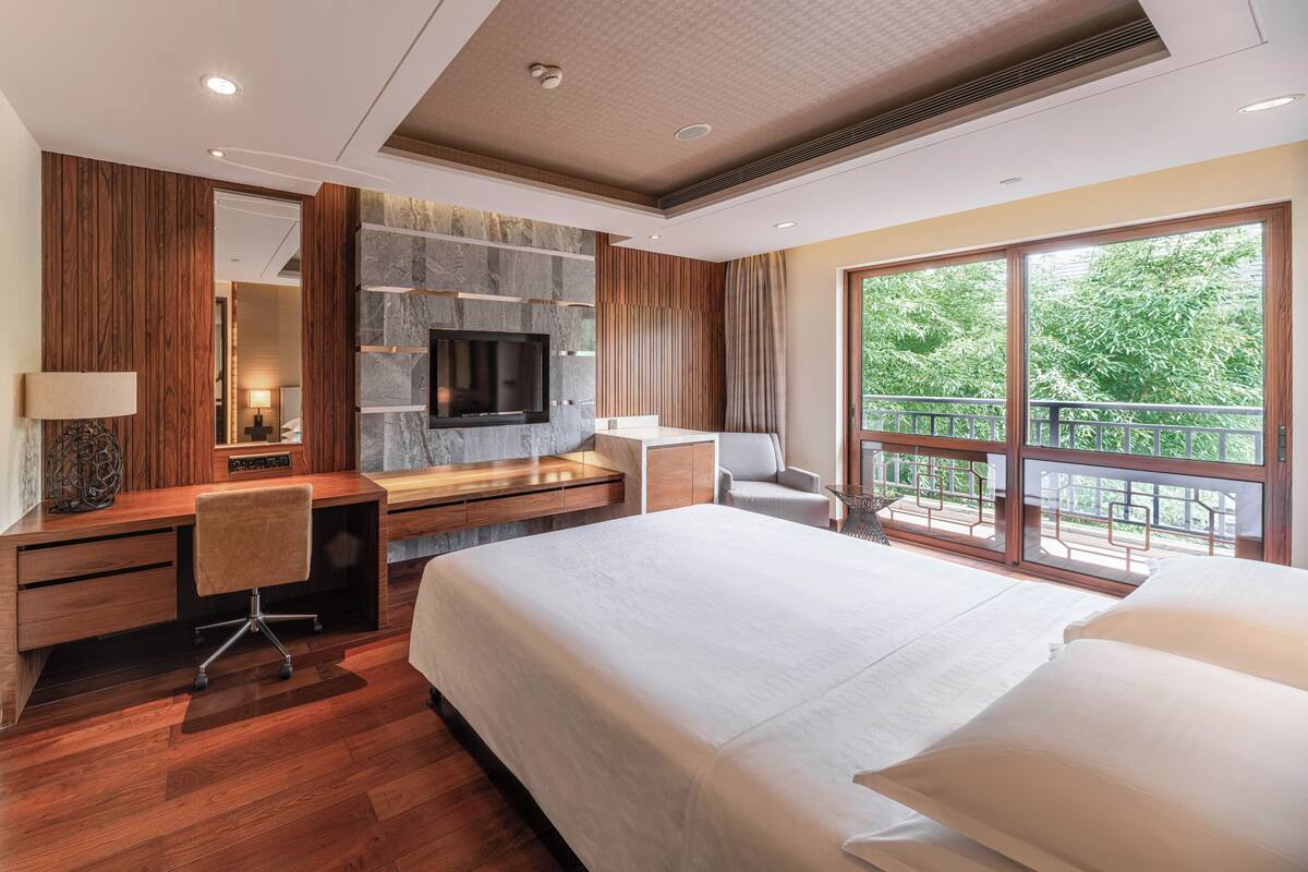 Una habitación de hotel moderna con una cama grande, un televisor montado en una pared de piedra, un escritorio con una silla y un balcón con vistas a zonas verdes.