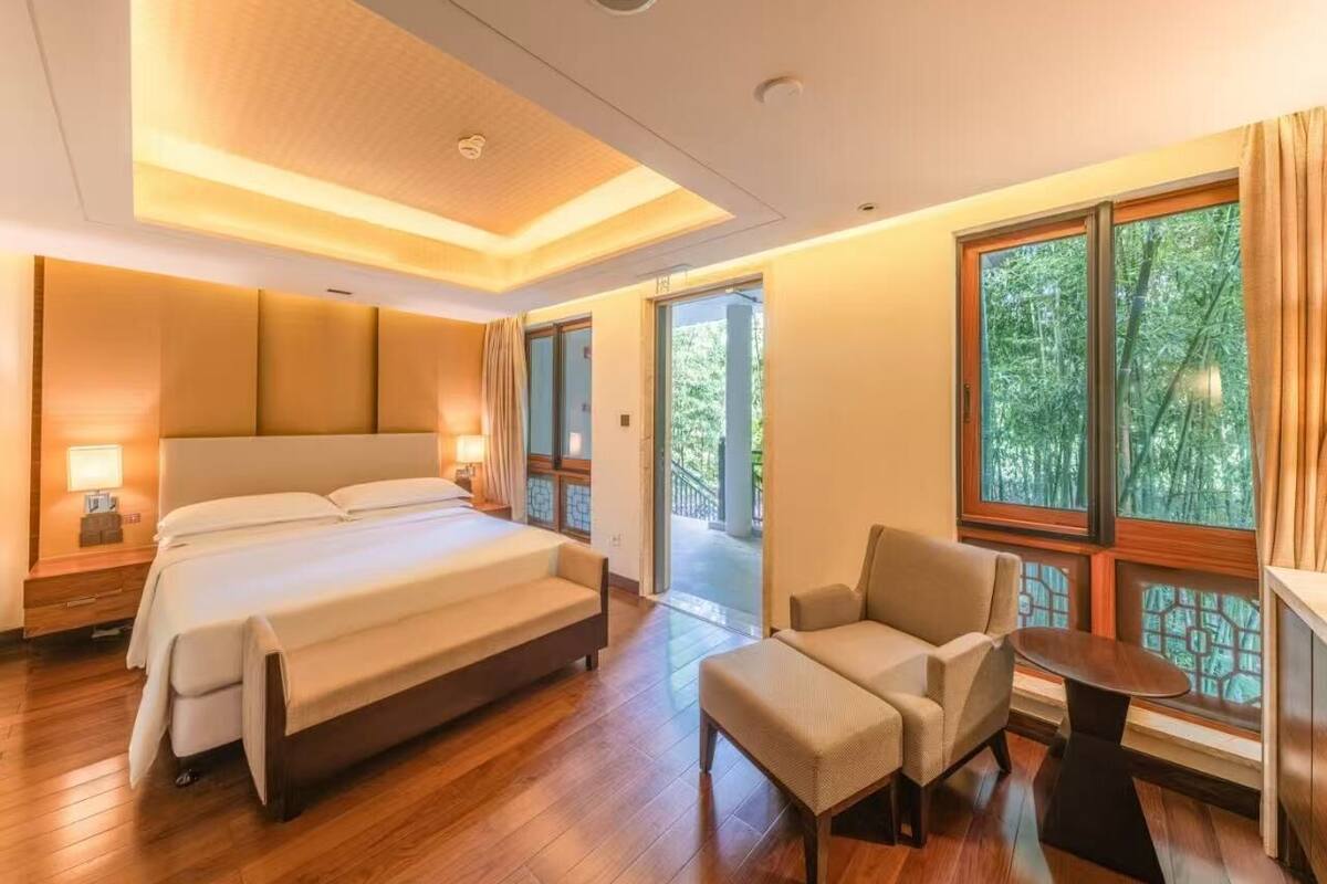 Habitación de hotel moderna con una cama grande, zona de estar y vistas a zonas verdes por las ventanas.