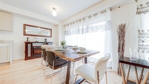 A dining area with a wooden table set for four, a kitchen in the background, and a large window with sheer curtains.