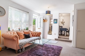 A living room with a tufted sofa, a glass coffee table, a purple rug, a dining area with a table and chairs, and a chandelier.