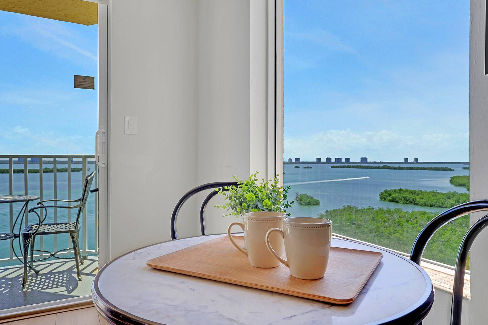 A balcony with a view of a bridge and water, a round table with two mugs, and a potted plant.