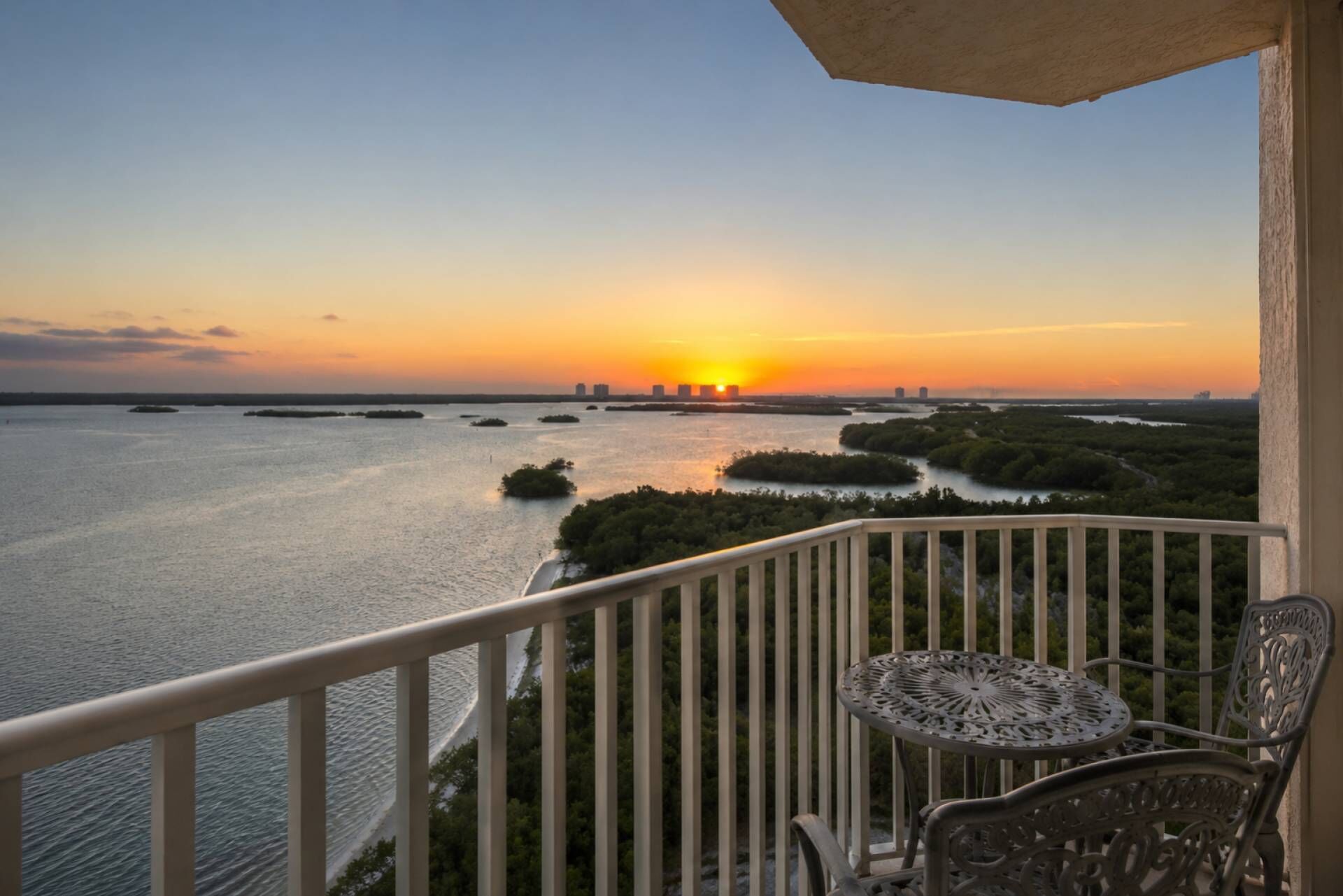 A balcony with a view of a sunset over a body of water, surrounded by greenery.